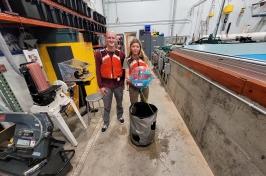 Two 主要研究 student researchers hold their winning ocean renewable energy device in front of a wave tank. 他们穿着红色的救生衣.